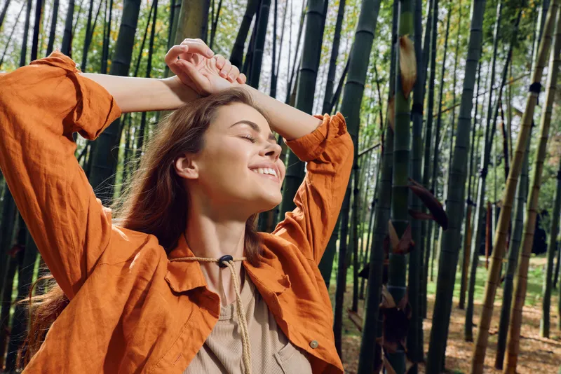 Woman smile nature bamboo forest outdoors relaxation portrait of a young female with eyes closed and arms raised, enjoying sunlight and fresh air in a serene woodland moment.