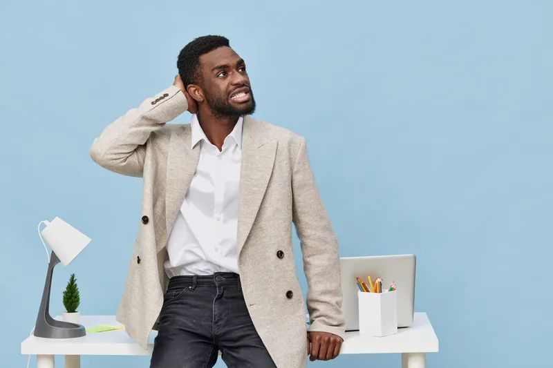 Smiling man with dark skin, curly hair, wearing a beige blazer and white shirt, standing in a modern office with a minimalist desk and blue background. People lifestyle concept, professional, casual, 