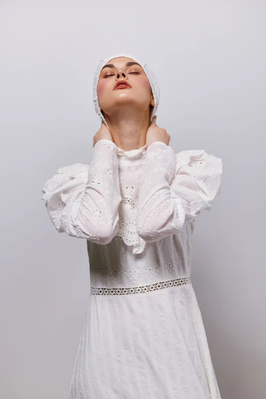 Elegant young woman in a white dress displaying serenity, with delicate puff sleeves and a minimalist background, embodying grace and simplicity in fashion