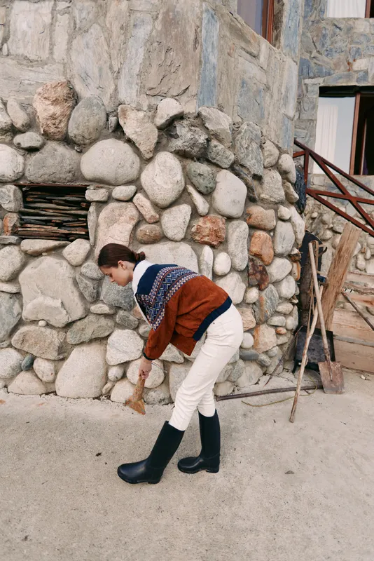 Woman stonewall sweater boots bending rural village portrait by a rustic building with stones, cozy knit layers and neutral brown tones for outdoor casual lifestyle and travel scene.