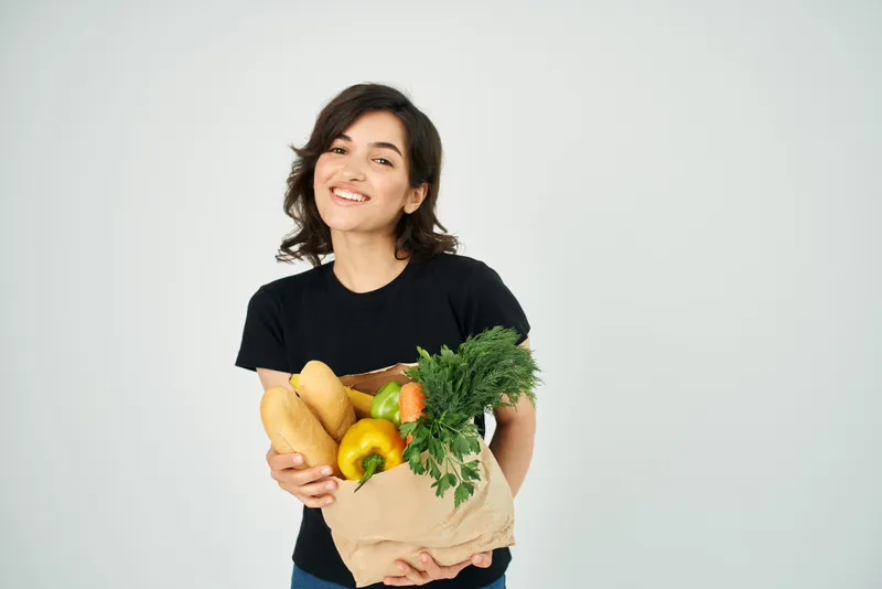 Веселая женщина с пакетом продуктов, овощами, здоровой пищей на светлом фоне