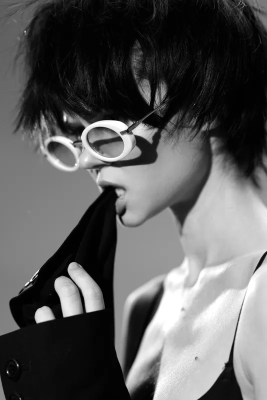 Black and white artistic portrait of a woman with short hair wearing round glasses biting a jacket sleeve.
