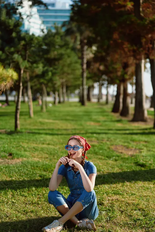 lifestyle woman in modern boho-western denim street style sitting relaxed on grass with sunglasses and red bandana, film color effect, sunny park background