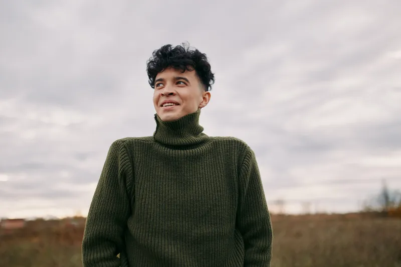 Portrait of a smiling young man in a cozy green sweater standing in an open field, conveying authenticity and veracity through candid expression and natural outdoor lighting, relaxed mood.