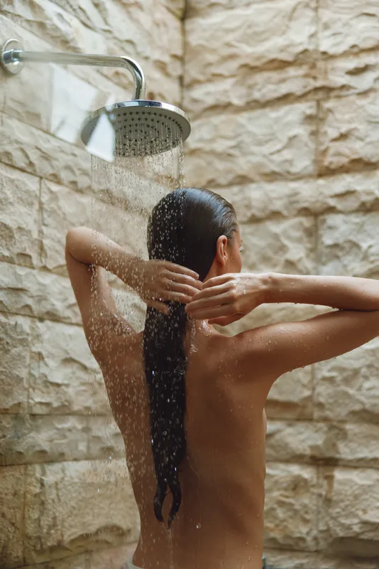 woman showering under rain shower head with wet hair, natural stone wall background, relaxed mood, warm tones, intimate bathroom scene