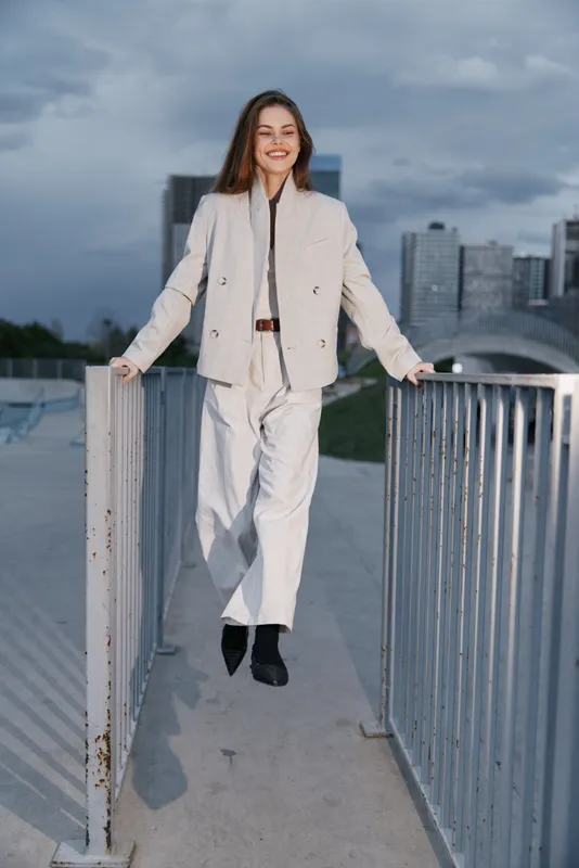 Confident woman in a light-colored business outfit smiling outdoors with a city skyline background, showcasing modern professionalism and style.