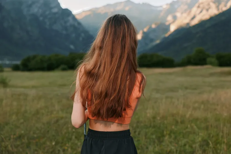 A person with long hair stands in a grassy meadow toward distant mountains, wearing an orange top and dark shorts. The scene evokes freedom, contemplation, and natural beauty.