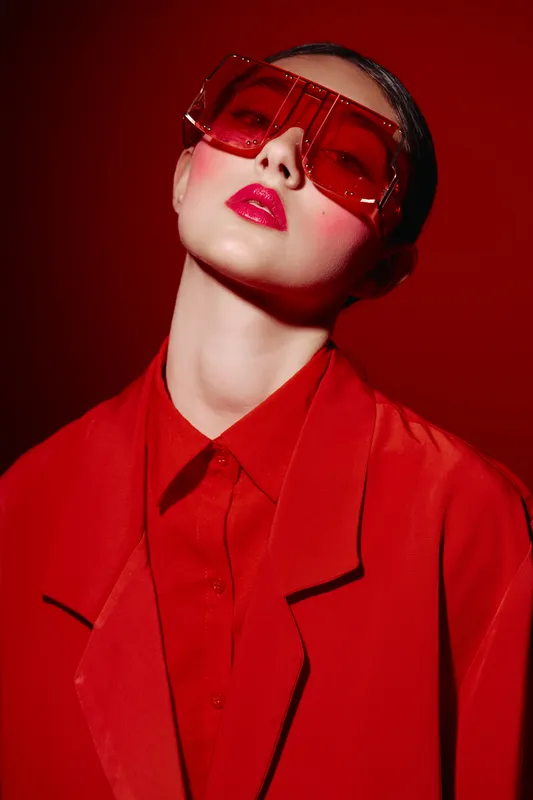 Fashion woman in stylish red oversized jacket with shiny lipstick and large square sunglasses posing on red background. Bold beauty, modern glamour, and creative fashion concept.