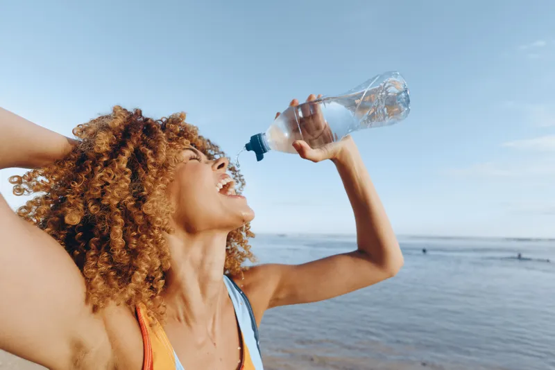 A young woman with curly hair on a sunny beach tilts her head back to drink water from a plastic bottle, enjoying a refreshing moment by the vast ocean and clear blue sky.