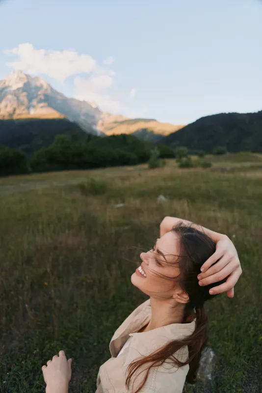 Joyful woman outdoors in a meadow, enjoying sunlight, laughter, and a gentle breeze with mountains in the distance, a serene lifestyle moment in nature