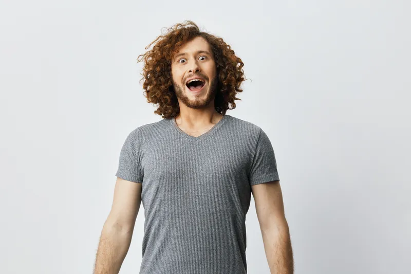 Happy young man with curly hair wearing a casual gray t shirt, expressing joy against a minimalistic background, perfect for lifestyle and happiness concepts