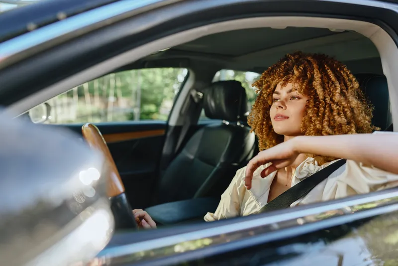 Young woman with curly hair driving a car, wearing seatbelt, relaxed and focused, natural daylight, urban background, safe driving concept.