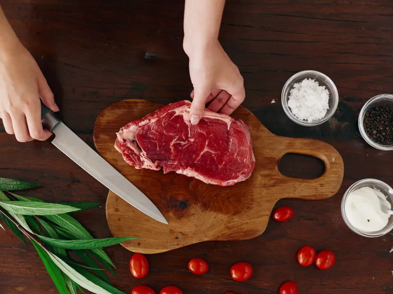 Женщина с ножом в руке нарезает свежее стейковое мясо для жарки на кухне
