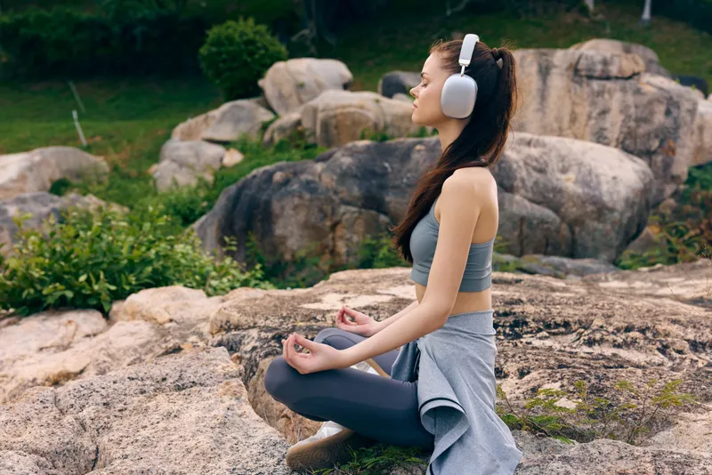 Young woman meditating on rocks in nature, wearing comfortable workout attire and headphones, promoting mindfulness and relaxation with a peaceful expression