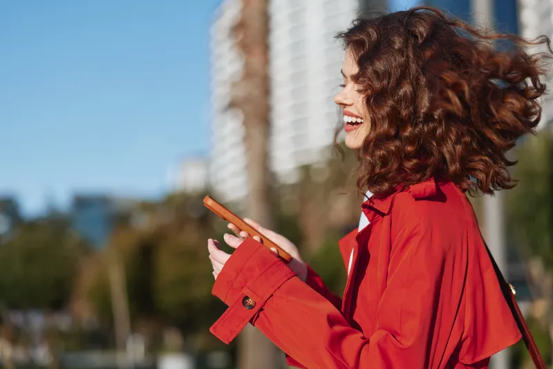 A smiling woman in a red jacket chats on her phone outdoors, wind in her hair, bright urban background, capturing casual street style and cheerful mood.