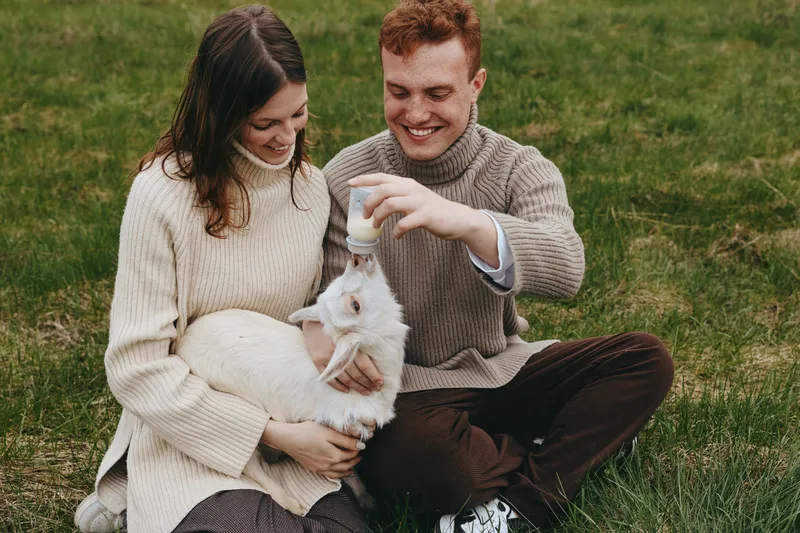 Couple outdoors with a goat kid, bottle feeding in a sunlit meadow, cozy sweaters, natural colors, relaxed lifestyle moment, candid farm atmosphere, joyful affection, simple rural scene