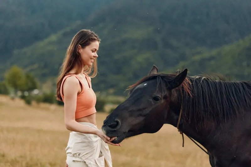 A cheerful woman in a coral top and light shorts stands in a grassy field, gently stroking a calm horse as hills rise in the distance, creating a serene outdoor moment.