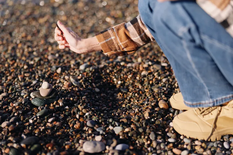Beach scene with a person crouching among rocks and stones, picking stones along the shoreline. A casual plaid shirt and jeans show outdoor exploration and a calm, mindful moment on the sand.