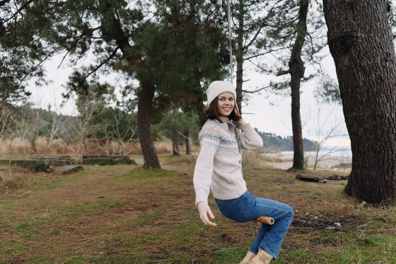 Woman in a knit sweater and beanie enjoys a playful moment outdoors among trees near a lakeside, wearing jeans and boots.