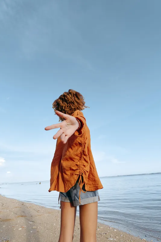 Curly woman wearing orange shirt and denim shorts standing on beach with arm extended, facing calm sea under clear blue sky. Concept of freedom, summer, and nature.