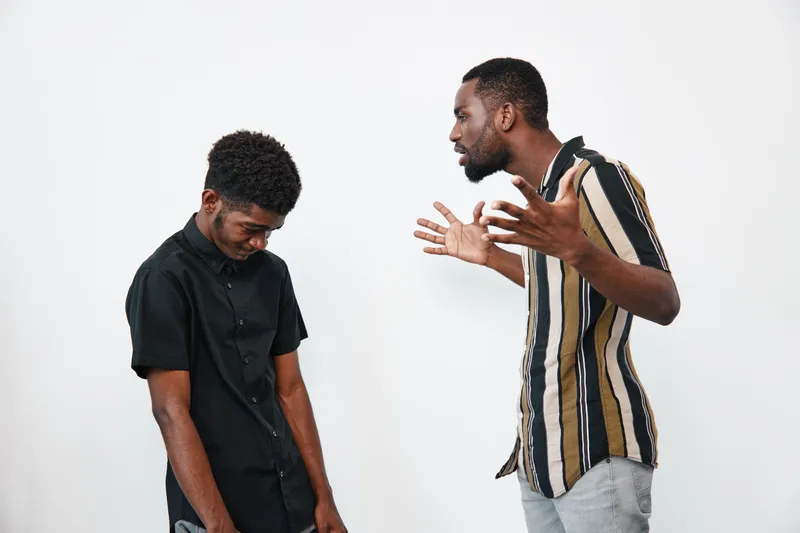 Two black African men with dark skin in casual clothes having a serious argument indoors on white background. One man is shouting while the other looks down in frustration.