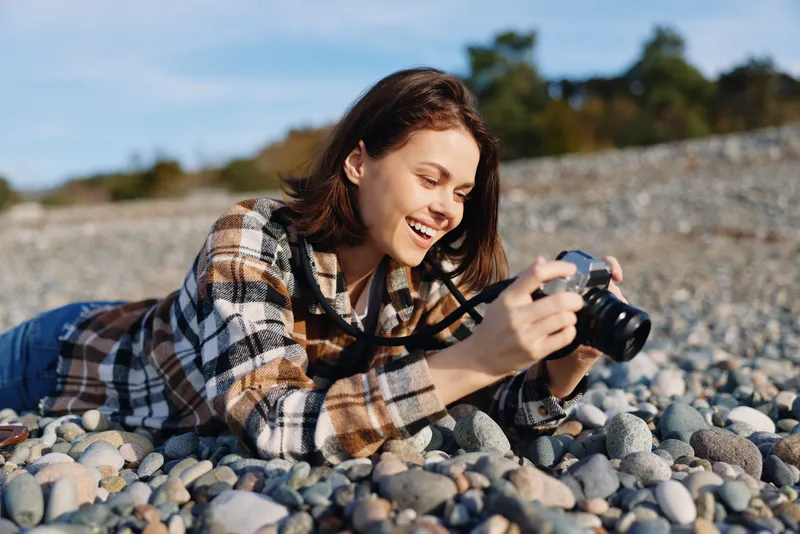 Photographer, woman lying on a rocky beach, smiling as she adjusts a camera, plaid jacket, sunlight on pebbles, capturing a candid outdoor moment with focus and expression and joy