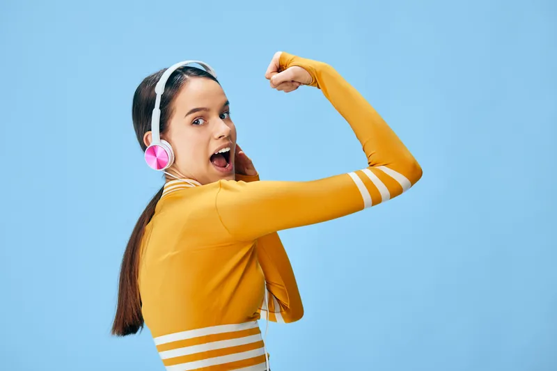Woman in orange athleticwear with headphones strikes a dynamic, powerful pose, showcasing fitness, sport energy and focused workout mindset against a bright blue backdrop.