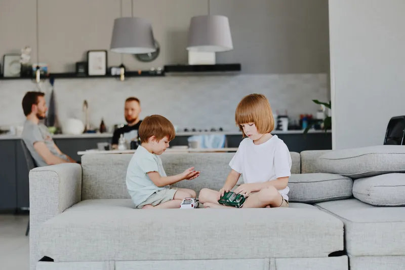 Two children playing together on a sofa while adults are relaxing in the background, showcasing a cozy family atmosphere and modern living space.