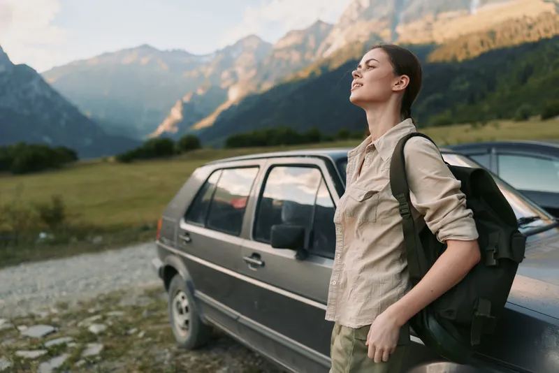 Young woman with backpack stands next to a car on a mountain road enjoying fresh air and nature during a hiking trip in the countryside.