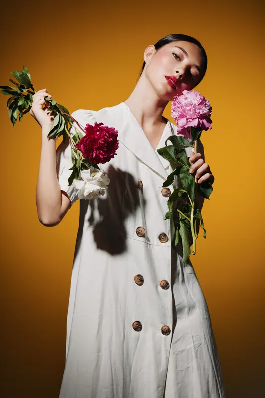 Fashion model wearing a white buttoned dress holds vibrant pink and red peonies against mustard background, expression calm and reflective. Concept for beauty, creative, and fashion.