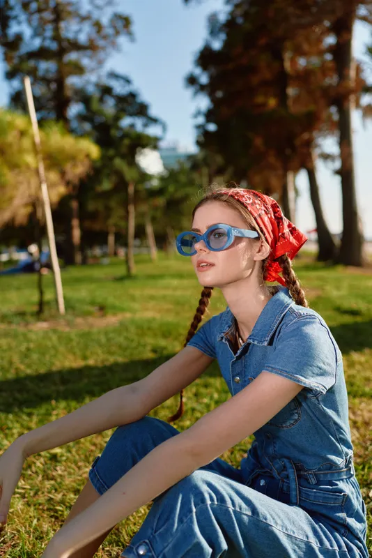 lifestyle woman in modern boho-western denim street style with blue sunglasses and red bandana, enjoying sunny outdoor park setting in film color tones