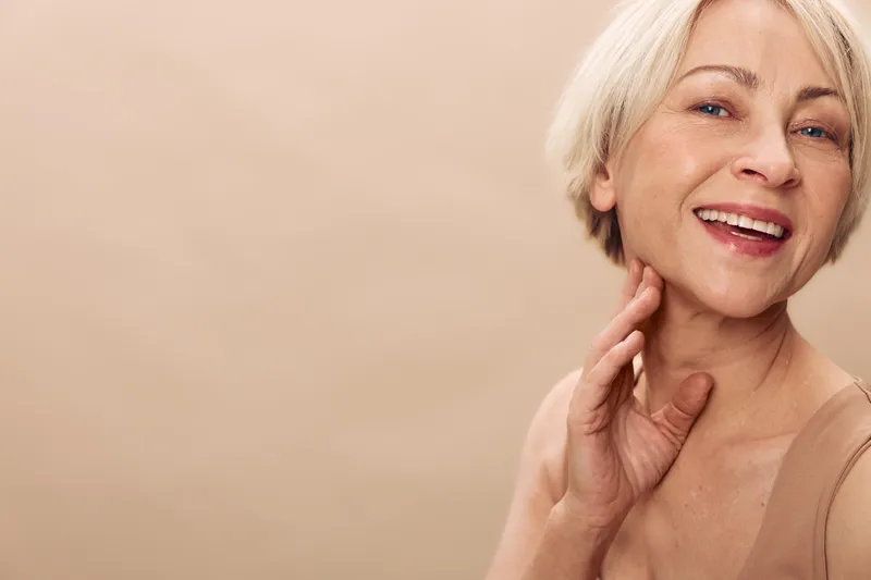 Smiling mature woman with short blonde hair touches neck gently, displaying natural beauty and confidence against soft beige background. Concept of aging gracefully and self-care.