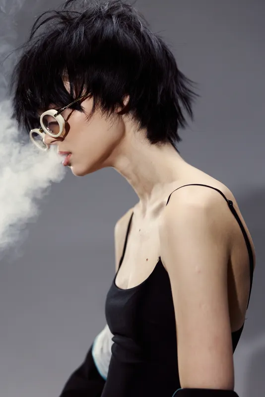Stylish woman with short black hair exhaling smoke, wearing fashionable sunglasses and a black top in a minimalistic studio setting.