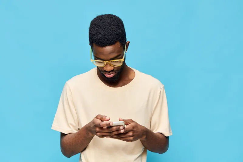 Young African man with glasses checking his phone against a bright blue background, wearing a casual beige t shirt Modern lifestyle concept