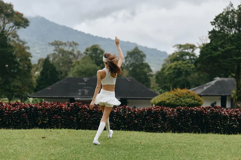 Young woman in a white crop top and skirt dancing joyfully outdoors in a green park with mountainous background, evoking happiness and freedom