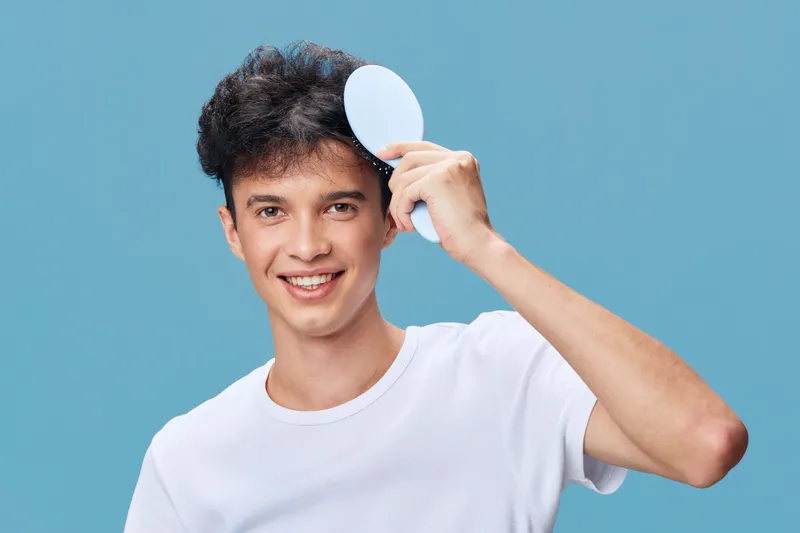 smiling man holding a mirror with black curly hair against a blue background wearing a white shirt expressing happiness and freshness