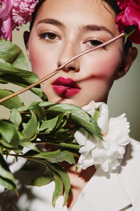 Close-up of a young woman with colorful pink and white flowers and vibrant green leaves around her face, wearing dark pink lipstick and soft fashion makeup in a creative beauty shot.