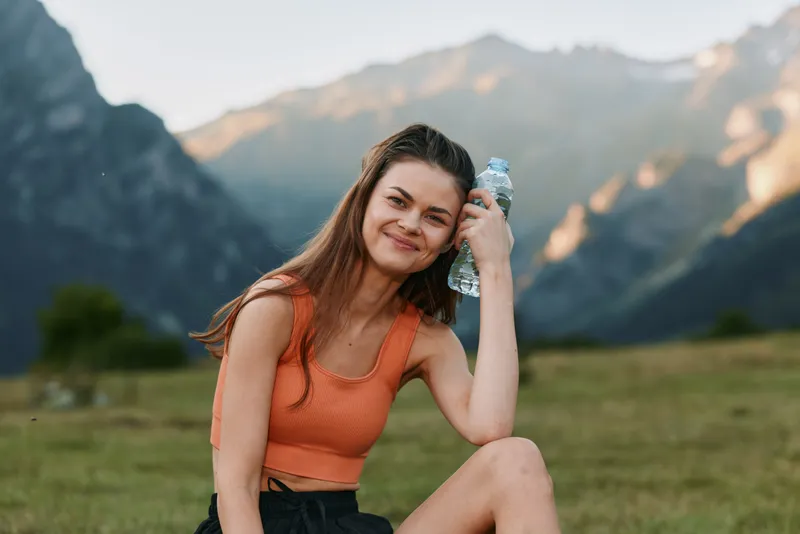 A smiling woman sits outdoors in a scenic mountain landscape, wearing an orange tank top and holding a water bottle, enjoying a sunny, relaxed moment in nature.