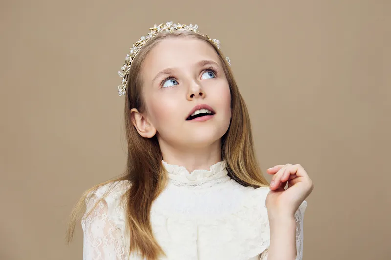 Young girl with long hair in a white dress and floral crown, looking up with a thoughtful expression against a neutral beige background