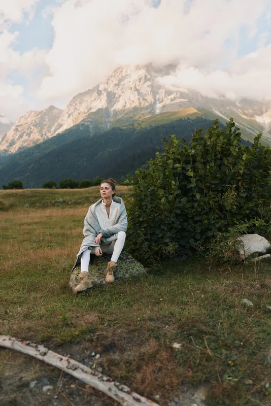 One person sits on a rock in a meadow, gazing thoughtfully at distant mountains, wrapped in a cozy cardigan for a quiet outdoor moment