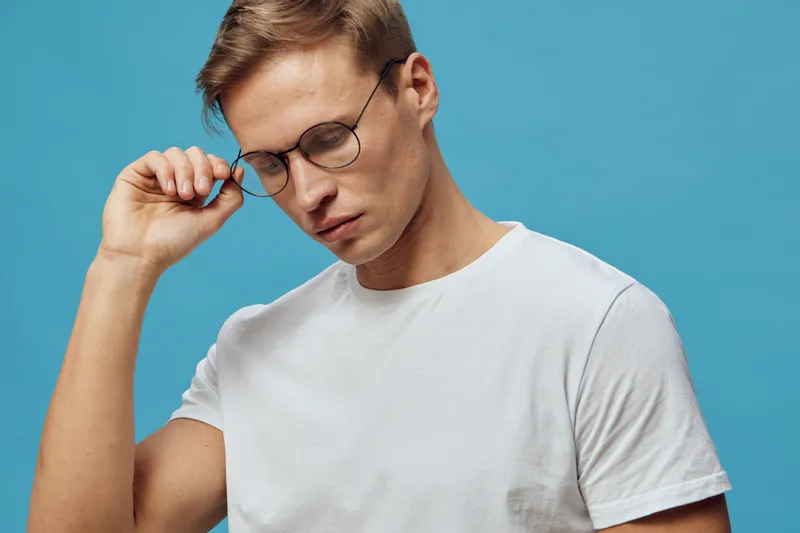 Thoughtful young man with glasses adjusting frame wearing plain white t-shirt look down isolated on blue background studio portrait. People lifestyle concept, introspection, concentration, fashion acc