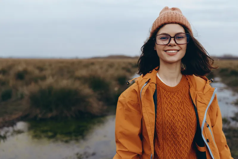 Woman lifestyle outdoors, smiling in autumn, wearing hat and glasses, orange coat, standing in nature landscape, soft wind, calm cloudy light, rural background, open field path