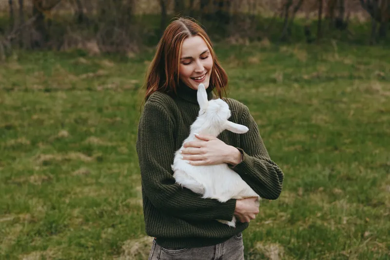 Young woman in a green sweater joyfully holds a white goat kid outdoors in a grassy field, enjoying a peaceful moment with nature in a casual lifestyle setting.