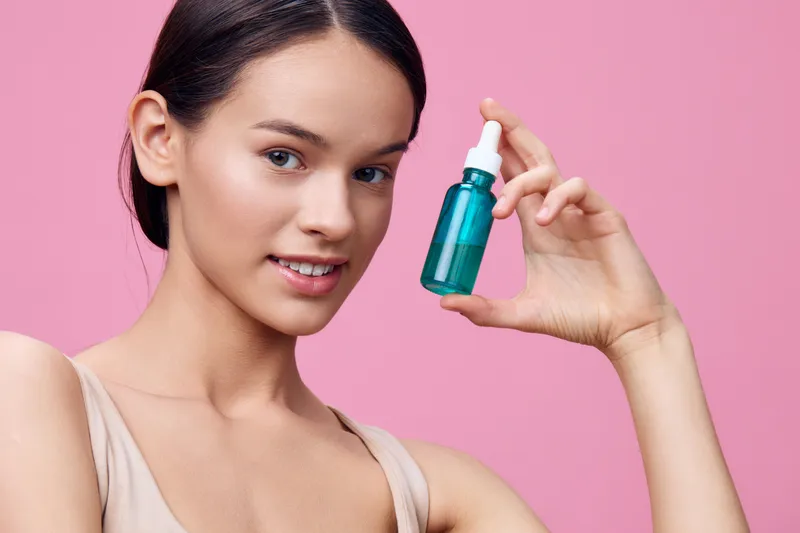 Young woman holding a blue serum bottle, smiling, against a pink background, showcasing beauty and skincare concepts, ideal for wellness and cosmetics themes