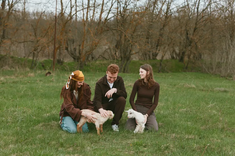 Lifestyle scene with three people in a field, interacting with small goats, calm joyful moment, casual clothing, natural light, countryside setting, three friends bonding with animals.