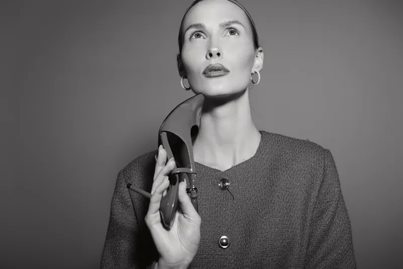 A fair skinned white woman in a textured coat holds a high heel shoe near her face, looking upward with a thoughtful expression. Studio portrait in black and white.