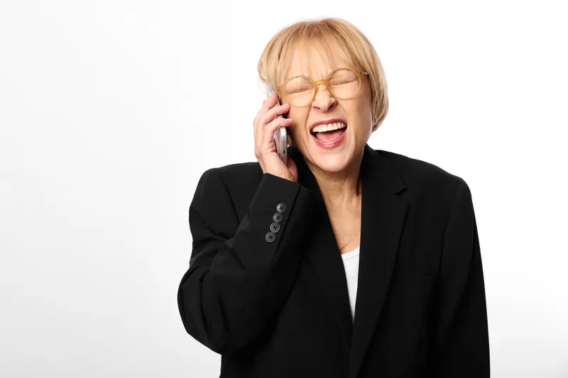 Cheerful woman in business suit talking on phone during a business call, office communication on white background, laughter and confident smile, friendly mood.