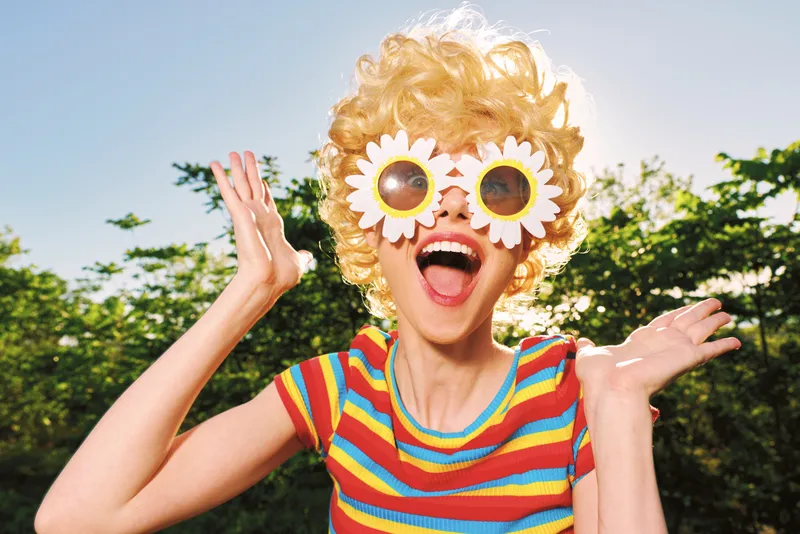 A joyful child in a bright striped shirt wears oversized sunflower sunglasses, arms raised in delight in a sunny outdoor setting. A playful moment capturing carefree summer energy.