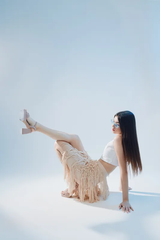 Young woman with long hair wearing sunglasses and a fringed skirt poses sitting on the floor in a bright studio. Concept of beauty fashion and summer style.