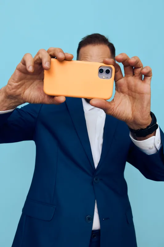 A confident business professional captures a selfie on a vibrant orange phone against a bright blue background, conveying energy, focus, and modern work life.